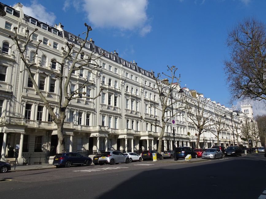 Photograph of a multi-storey white terraced building with ornate architectural details, including decorative cornices and window moldings, situated on Belgrave Square in Belgravia. The facade features