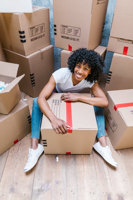 Two women standing inside a room surrounded by stacked cardboard moving boxes, some sealed with red packing tape. One woman is holding a box with her left hand, and the other woman is holding a large